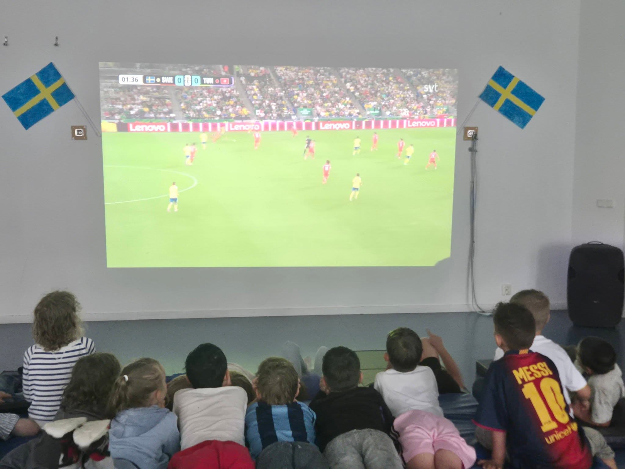 Barn sitter på golvet och tittar på en fotbollslandskamp på storbild mellan två svenska flaggor.