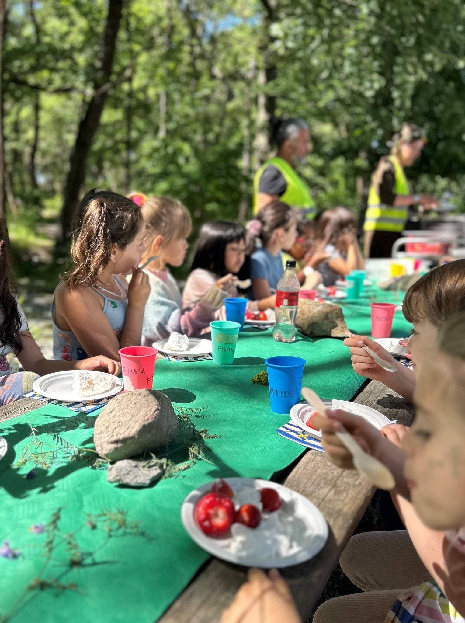Fritidsbarn äter vid ett dukat bord i skogen medan pedagoger grillar.