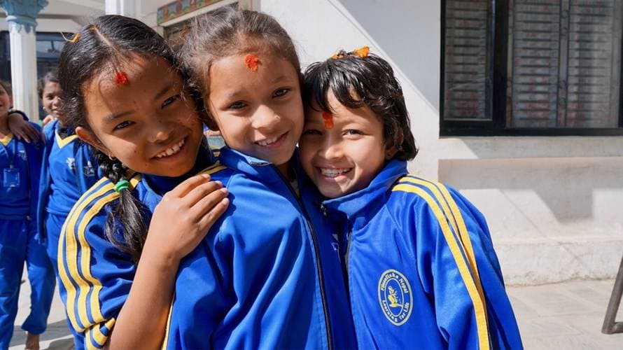Tre flickor i blå overaller med tika på pannan kramas leende på skolgården i Nepal.