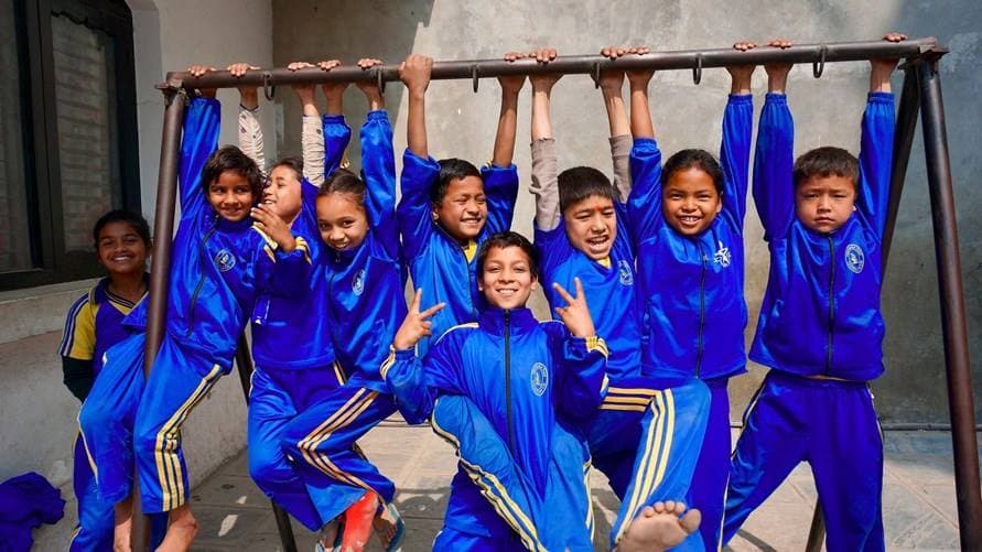 Åtta elever i blå overaller hänger glada på en gympaställning utomhus i Nepal.