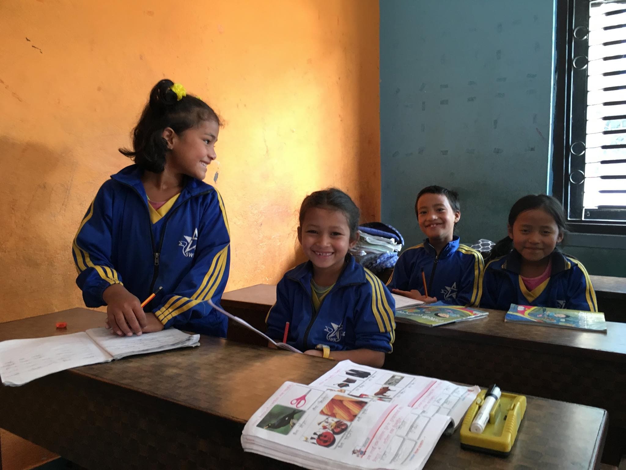 Fyra leende elever i blå skoluniformer vid trä-skolbänkar i ett klassrum i Nepal.
