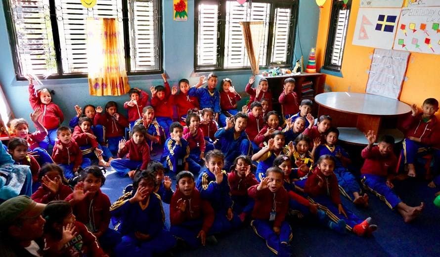 Stor klass med elever på golvet vinkar mot kameran i ett ljust klassrum i Nepal.